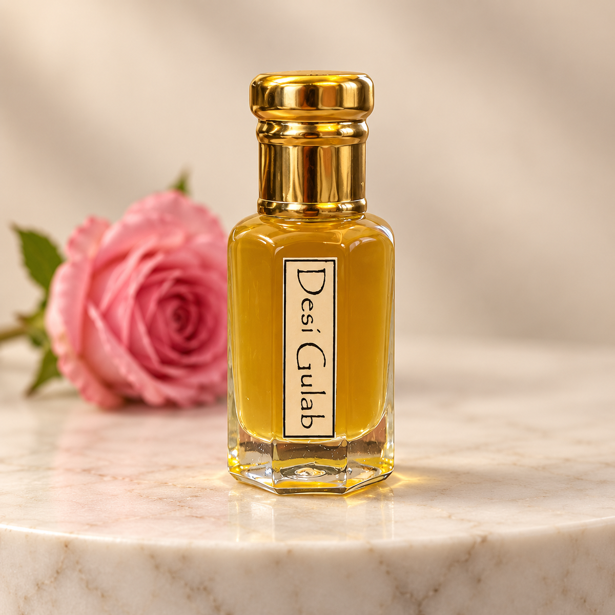 A small rectangular glass perfume bottle filled with golden-yellow liquid, topped with a shiny gold cap, standing on a white marble surface. A pink rose is blurred in the background.
