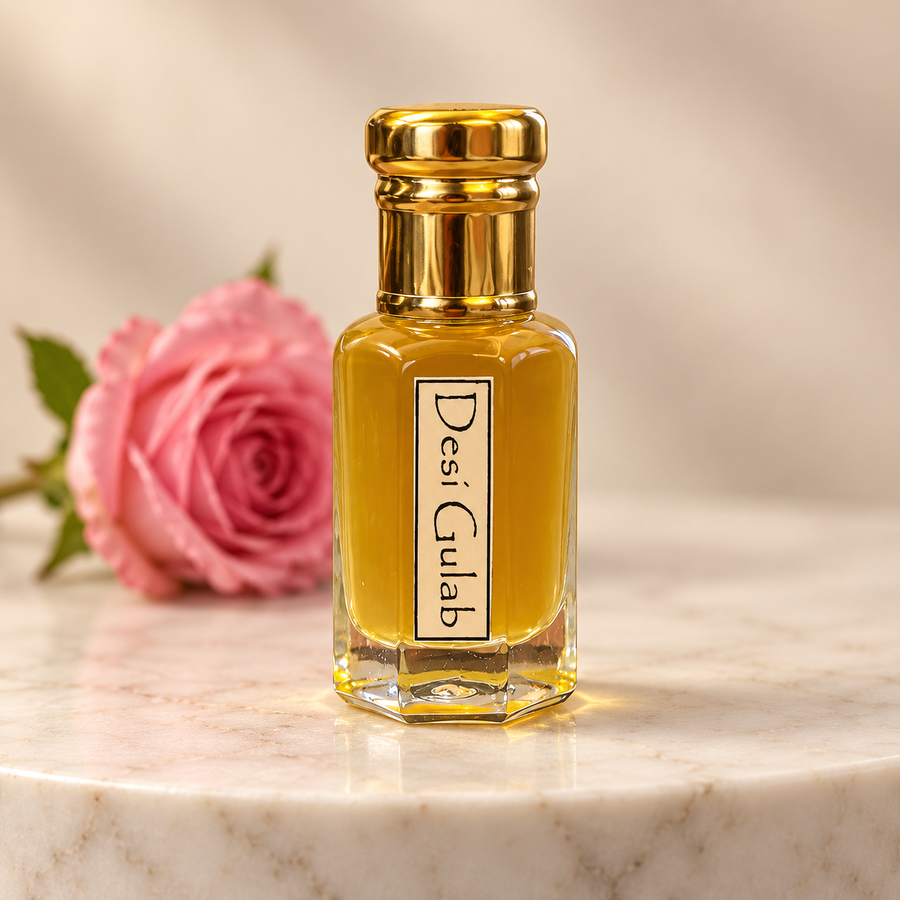 A small rectangular glass perfume bottle filled with golden-yellow liquid, topped with a shiny gold cap, standing on a white marble surface. A pink rose is blurred in the background.