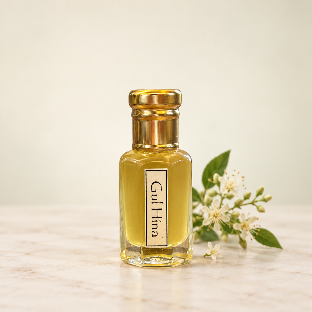 A small clear glass bottle filled with golden yellow liquid, topped with a metallic gold cap, and labeled 'Gul Hina.' It sits on a light marble surface with white flowers and green leaves beside it against a soft cream background.